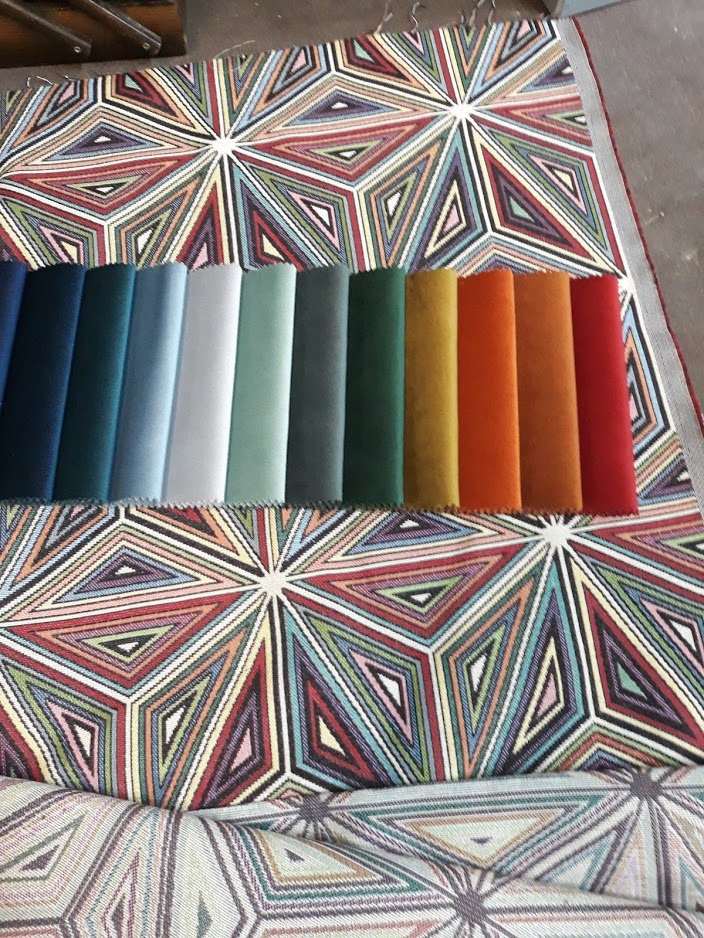 Próbniki tkanin tapicerskich w odcieniach niebieskiego, zieleni, brązu i czerwieni na tle tkaniny w geometryczny wzór.