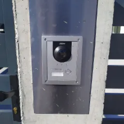 Srebrny panel wideodomofonu Dahua zamontowany na metalowej płycie na betonowym słupku bramy wjazdowej.