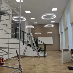 Wnętrze komercyjne w trakcie instalacji oświetlenia: widoczne okrągłe lampy LED, rusztowanie, drabiny, schody z barierką, podłoga imitująca drewno, biała tablica na ścianie.
