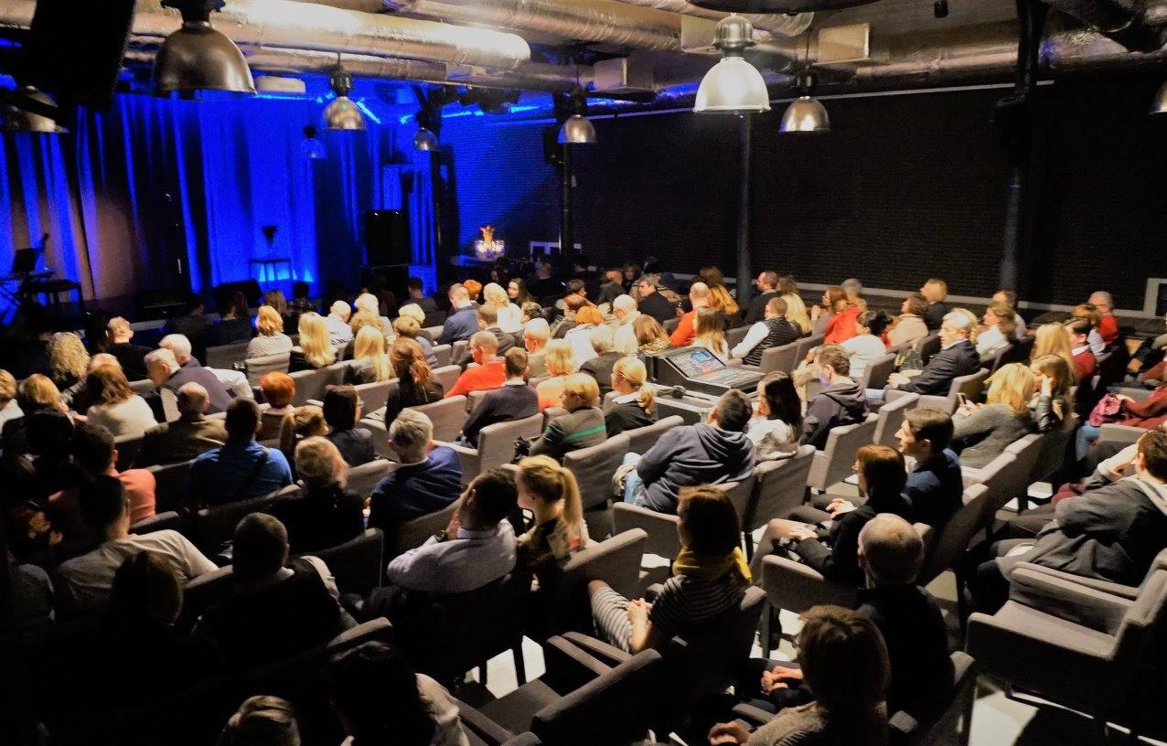 Widok z tyłu na publiczność siedzącą w rzędach krzeseł w sali widowiskowej w Szczecinie, z oświetloną sceną w tle.