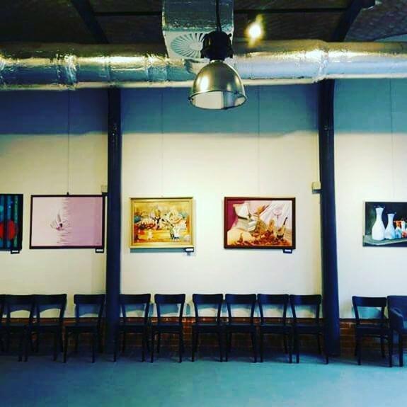 Sala z rzędem krzeseł przed ścianą z wiszącymi obrazami w ramach, oświetlona metalową lampą wiszącą i widocznymi elementami wentylacji sufitowej.