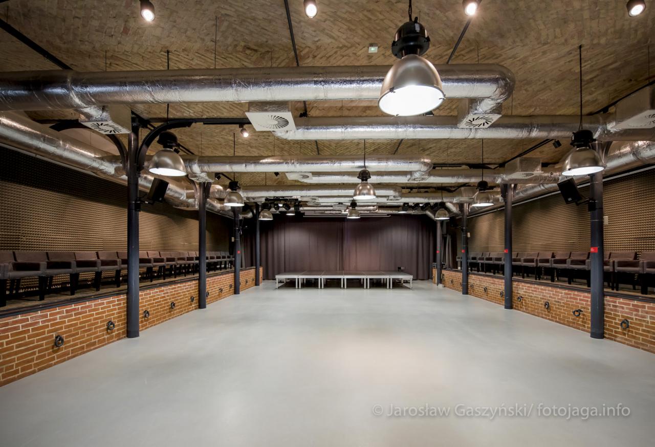 Przestronna sala widowiskowa z ceglanymi ścianami, rzędami krzeseł na podwyższeniu, sceną z brązowymi zasłonami i surowym sufitem z widocznymi rurami wentylacyjnymi oraz metalowymi lampami.