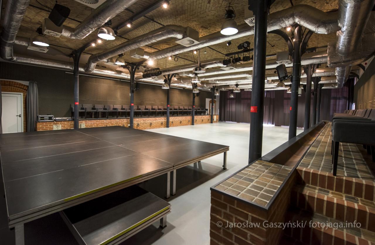 Duża sala eventowa z podwyższoną sceną, rzędami krzeseł w tle, surowym ceglanym wykończeniem i widocznymi instalacjami wentylacyjnymi pod sufitem.