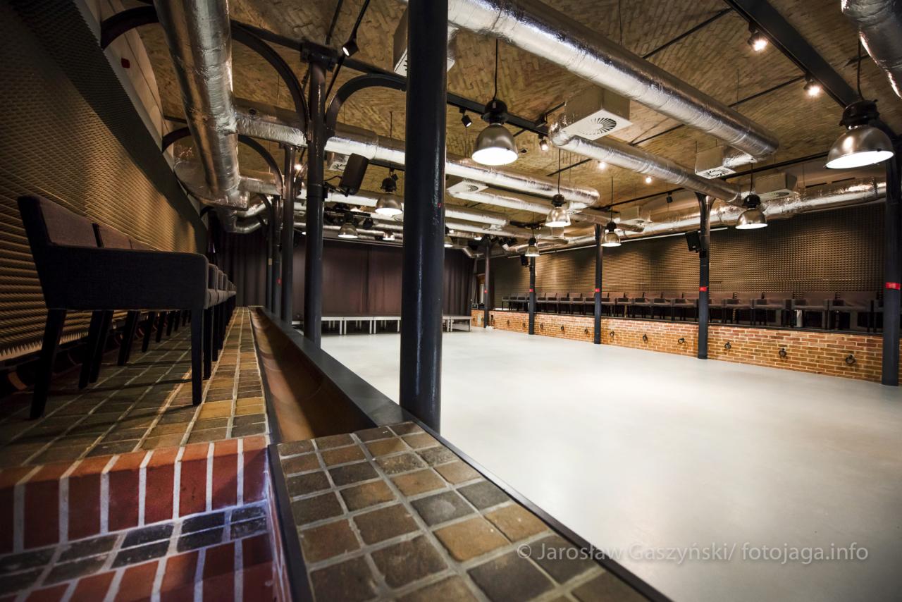 Przestronna sala widowiskowa z ceglanymi elementami, rzędami krzeseł i widocznymi instalacjami wentylacyjnymi pod sufitem, przygotowana na wydarzenie.