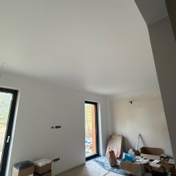 Luk Complex - Wnętrze po remoncie: białe ściany, okna z czarnymi ramami, podłoga zabezpieczona folią, kartony i materiały budowlane. Widok na zewnątrz przez okno.