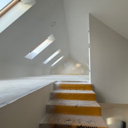 Luk Complex - Poddasze po remoncie z oknami dachowymi i schodami zabezpieczonymi folią ochronną. Ściany i sufit pomalowane na biało. Widoczne przygotowania do dalszych prac.