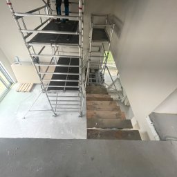 Luk Complex - Wysoka drabina rusztowaniowa ustawiona przy ścianie w trakcie prac wykończeniowych, widoczne ślady po szpachlowaniu, w tle okno i paleta z płytami.