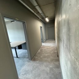 Luk Complex - Korytarz biurowy w szarej kolorystyce, z widoczną surową fakturą ściany, oświetleniem ledowym i otwartymi drzwiami do pomieszczenia z biurkiem.