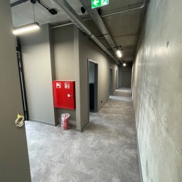 Luk Complex - Długi korytarz biurowy z szarą posadzką i ścianami, widoczna instalacja pod sufitem, czerwona szafka hydrantowa z gaśnicą obok, znak ewakuacyjny.