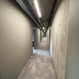Luk Complex - Długi korytarz biurowy z szarymi ścianami, betonowym sufitem z widocznymi rurami i oświetleniem liniowym, oraz szarą podłogą z płytek. Jedna ściana gładka, druga z surowego betonu.