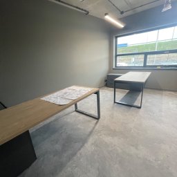 Luk Complex - Surowe, szare wnętrze biurowe z dwoma biurkami o metalowych nogach i drewnianych blatach. Na jednym z biurek leży biała tkanina ochronna. Widoczne szpachlowane ściany i betonowa podłoga.