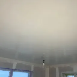 Wnętrze pokoju w trakcie prac wykończeniowych; widoczny gładki, jasny sufit z wiszącą żarówką oraz ściany z płyt kartonowo-gipsowych, częściowo przygotowane do dalszych prac.