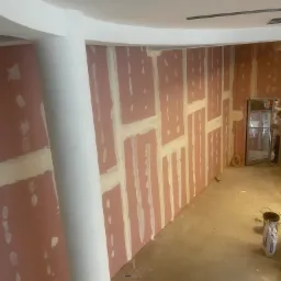 Montaż płyt kartonowo-gipsowych na ścianie w pomieszczeniu z zaokrąglonym sufitem, widoczne łączenia płyt wypełnione masą szpachlową, surowy beton na podłodze, w tle otwarte drzwi i wiadro z zaprawą.