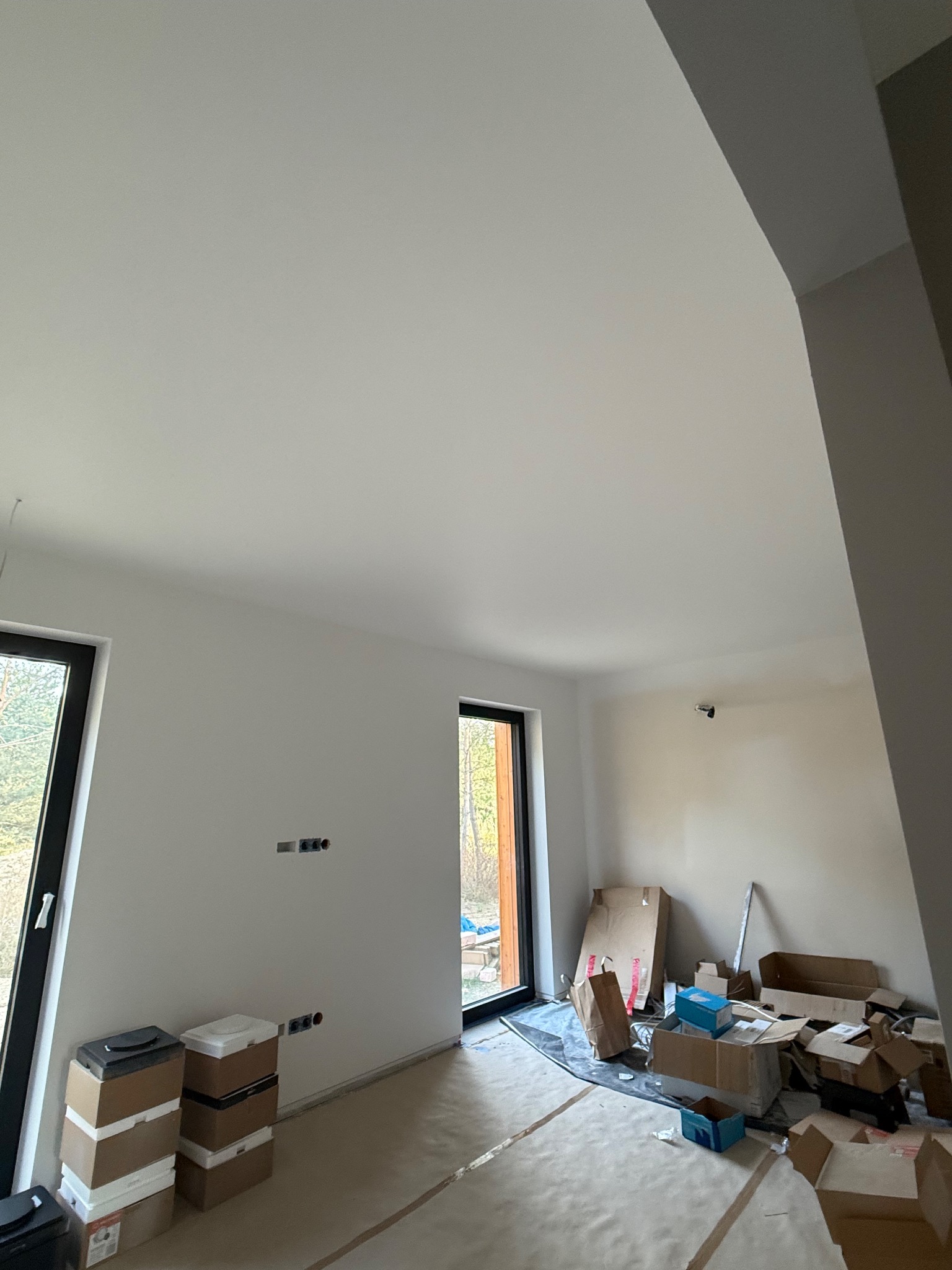 Wnętrze po remoncie: białe ściany, okna z czarnymi ramami, podłoga zabezpieczona folią, kartony i materiały budowlane. Widok na zewnątrz przez okno.