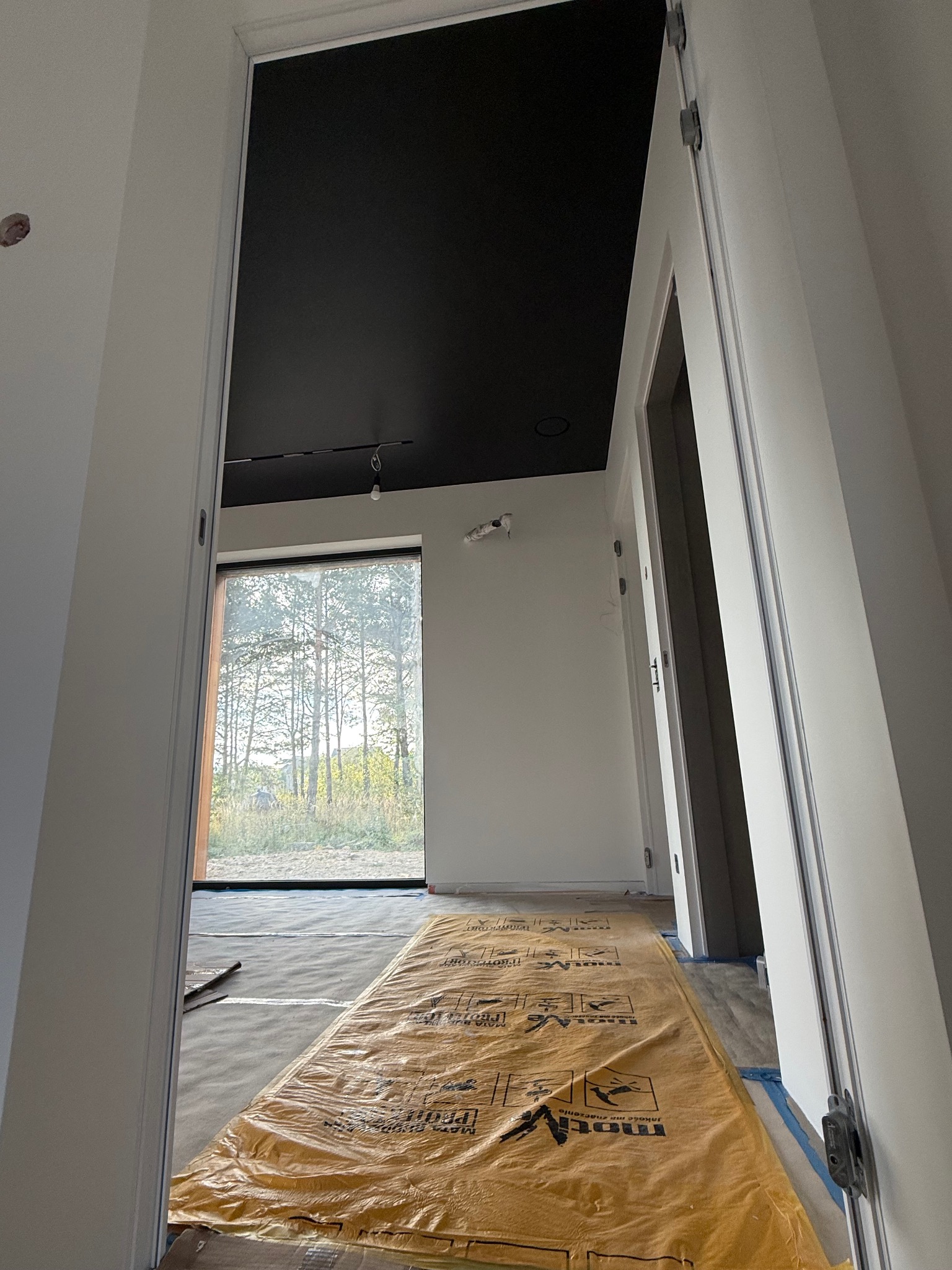 Wnętrze w trakcie prac wykończeniowych: czarny sufit, białe ściany, okno z widokiem na drzewa, podłoga zabezpieczona folią ochronną. Widok z perspektywy wejścia.