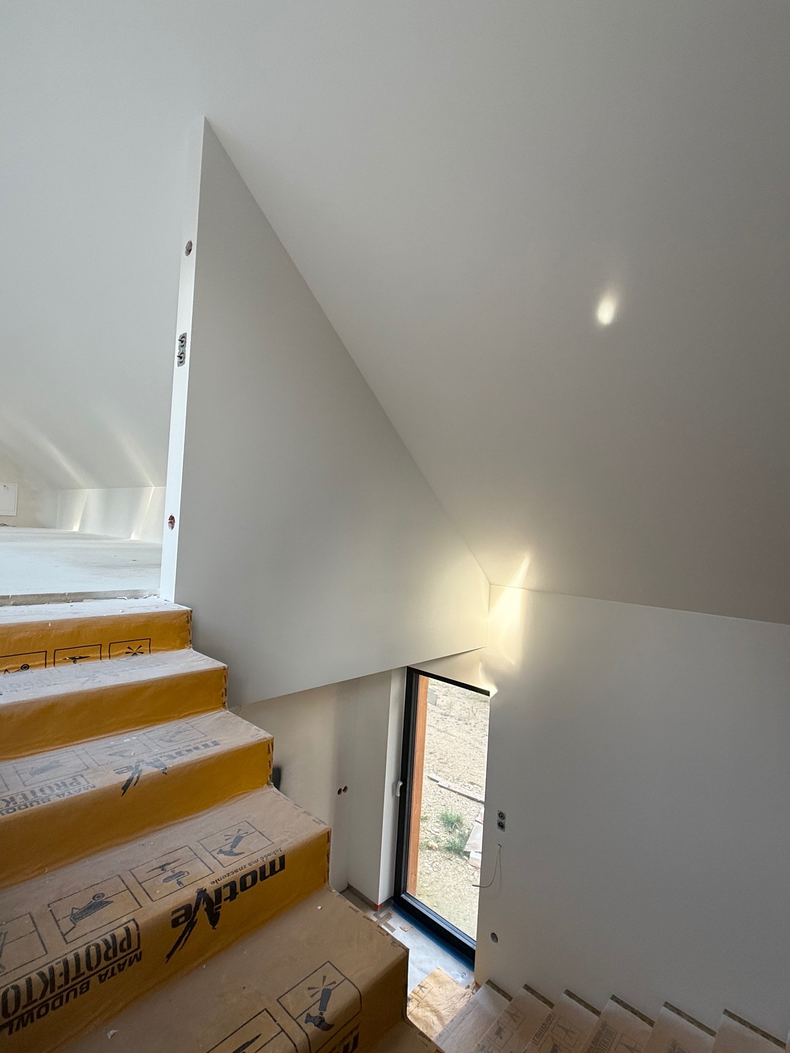 Białe, wygładzone ściany poddasza z oknem i schodami zabezpieczonymi kartonem budowlanym. Widoczne przygotowania do dalszych prac wykończeniowych.