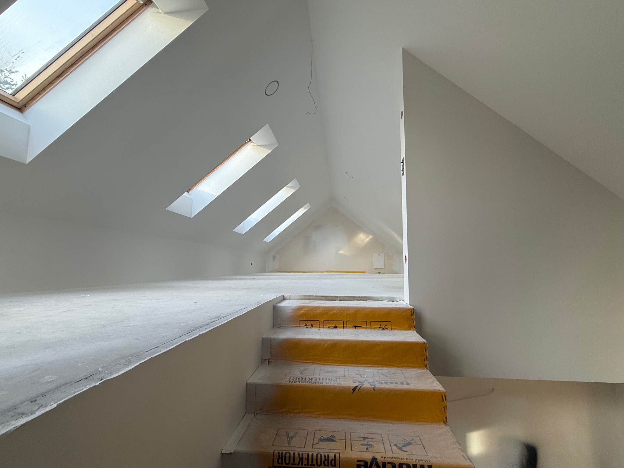 Poddasze po remoncie z oknami dachowymi i schodami zabezpieczonymi folią ochronną. Ściany i sufit pomalowane na biało. Widoczne przygotowania do dalszych prac.