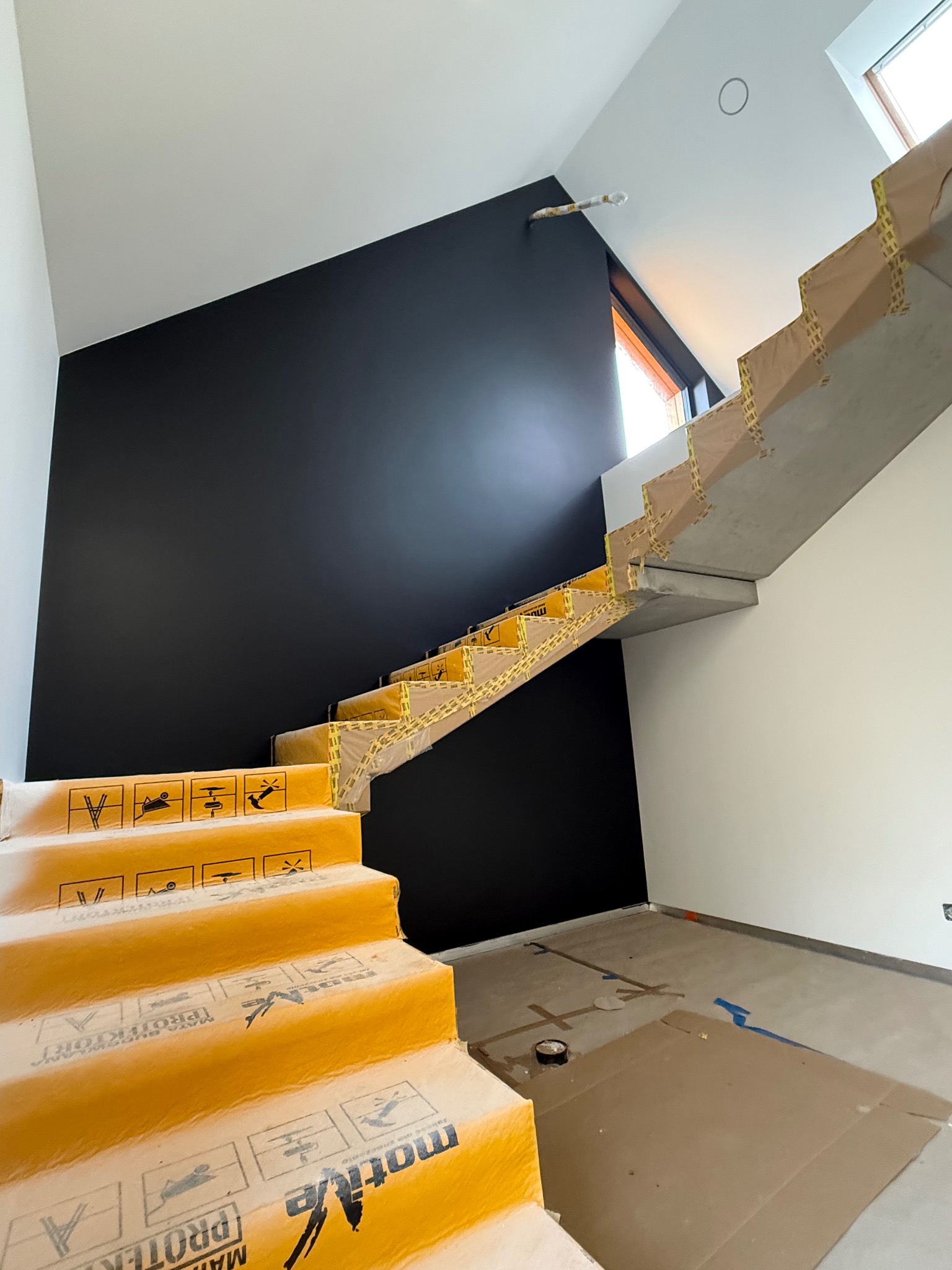 Betonowe schody zabezpieczone folią ochronną na czas prac wykończeniowych, czarna ściana w tle, surowe wnętrze w trakcie remontu.