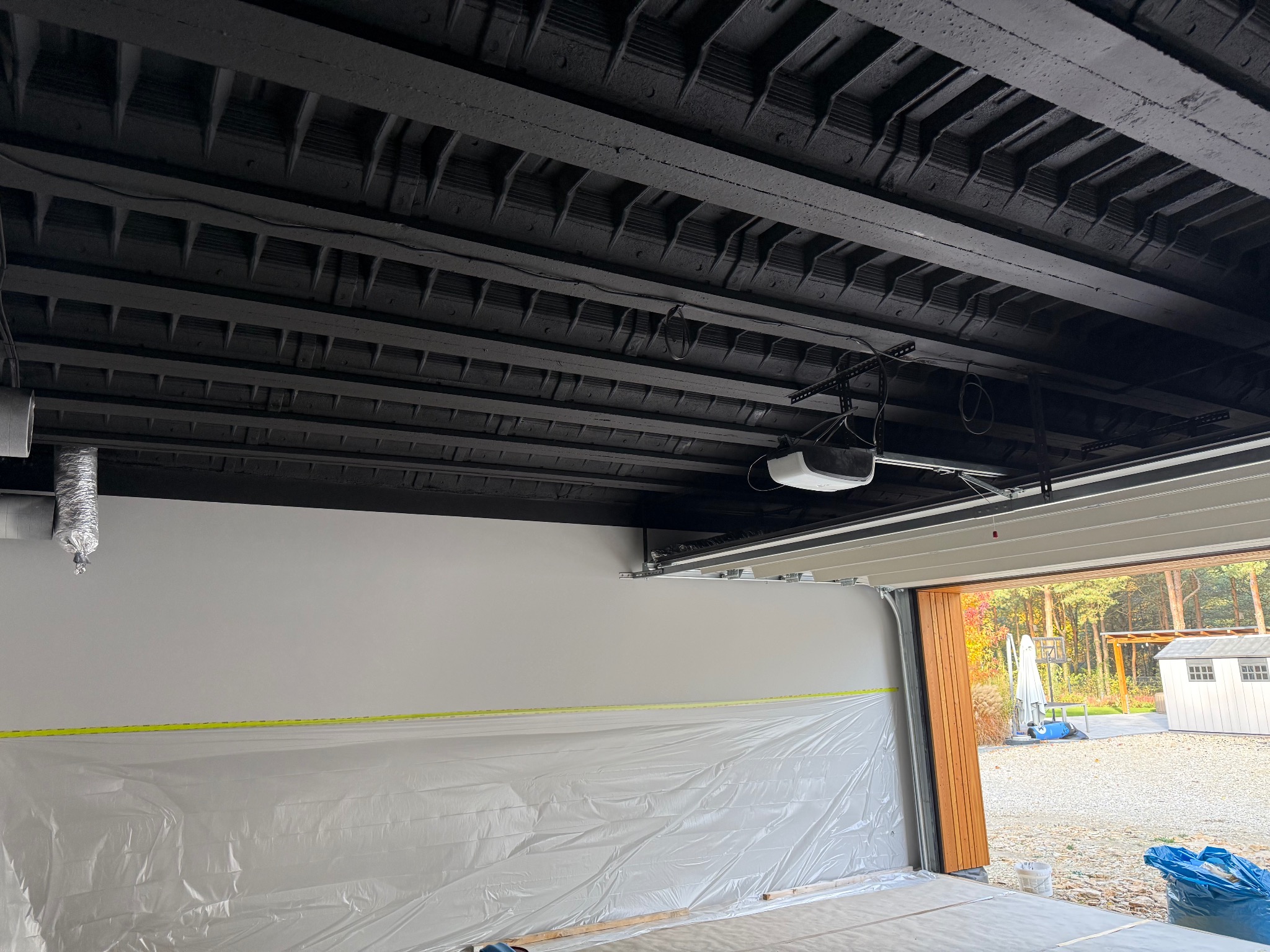 Pomieszczenie garażowe w trakcie prac malarskich: czarny sufit z belkami, białe ściany zabezpieczone folią malarską, otwarta brama garażowa z widokiem na zewnątrz.
