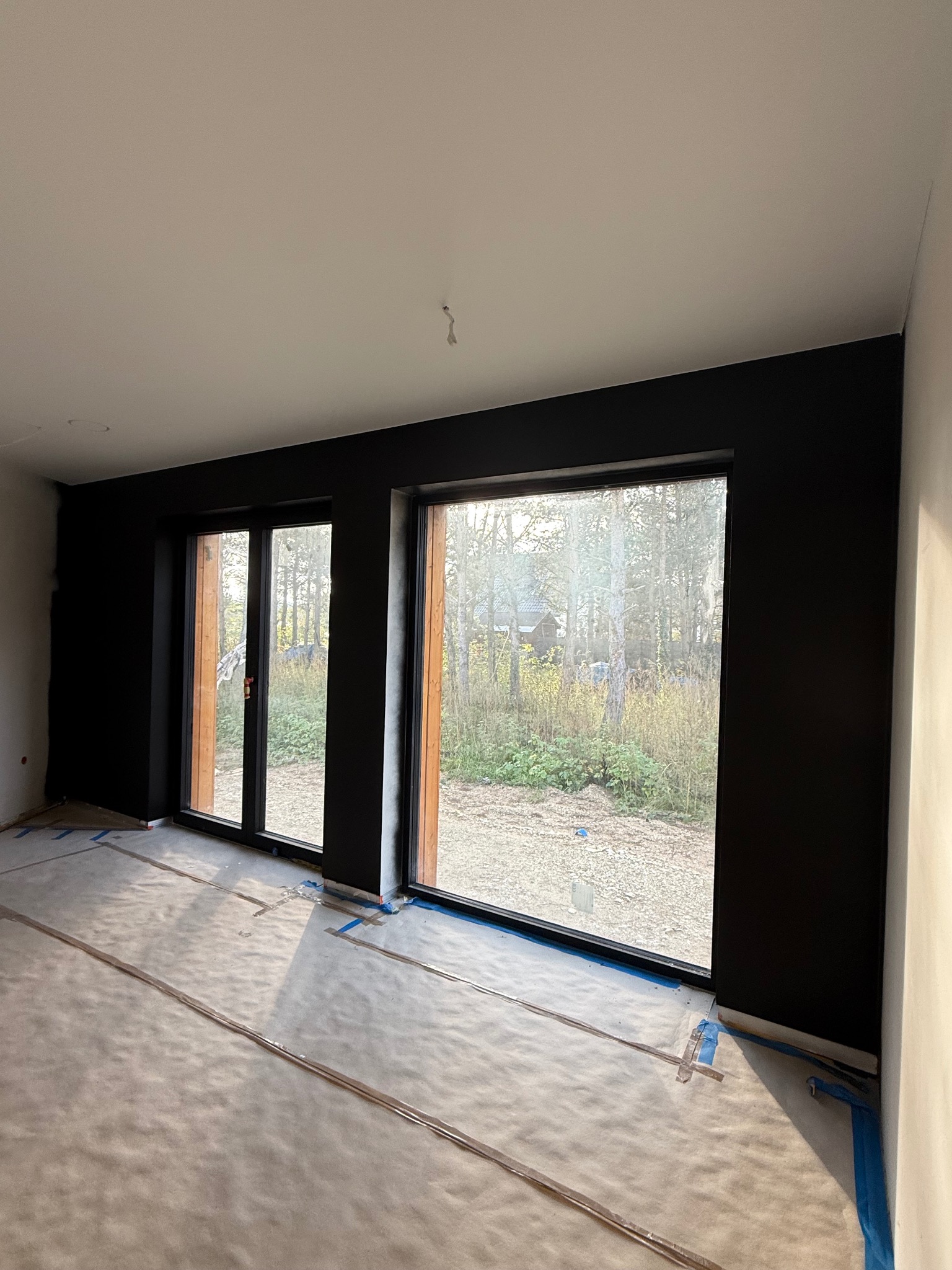 Pokój po malowaniu na czarno, zabezpieczona podłoga folią i taśmą. Widoczne duże okna z widokiem na zewnątrz. Ściany pomalowane na jednolity kolor, sufit biały.