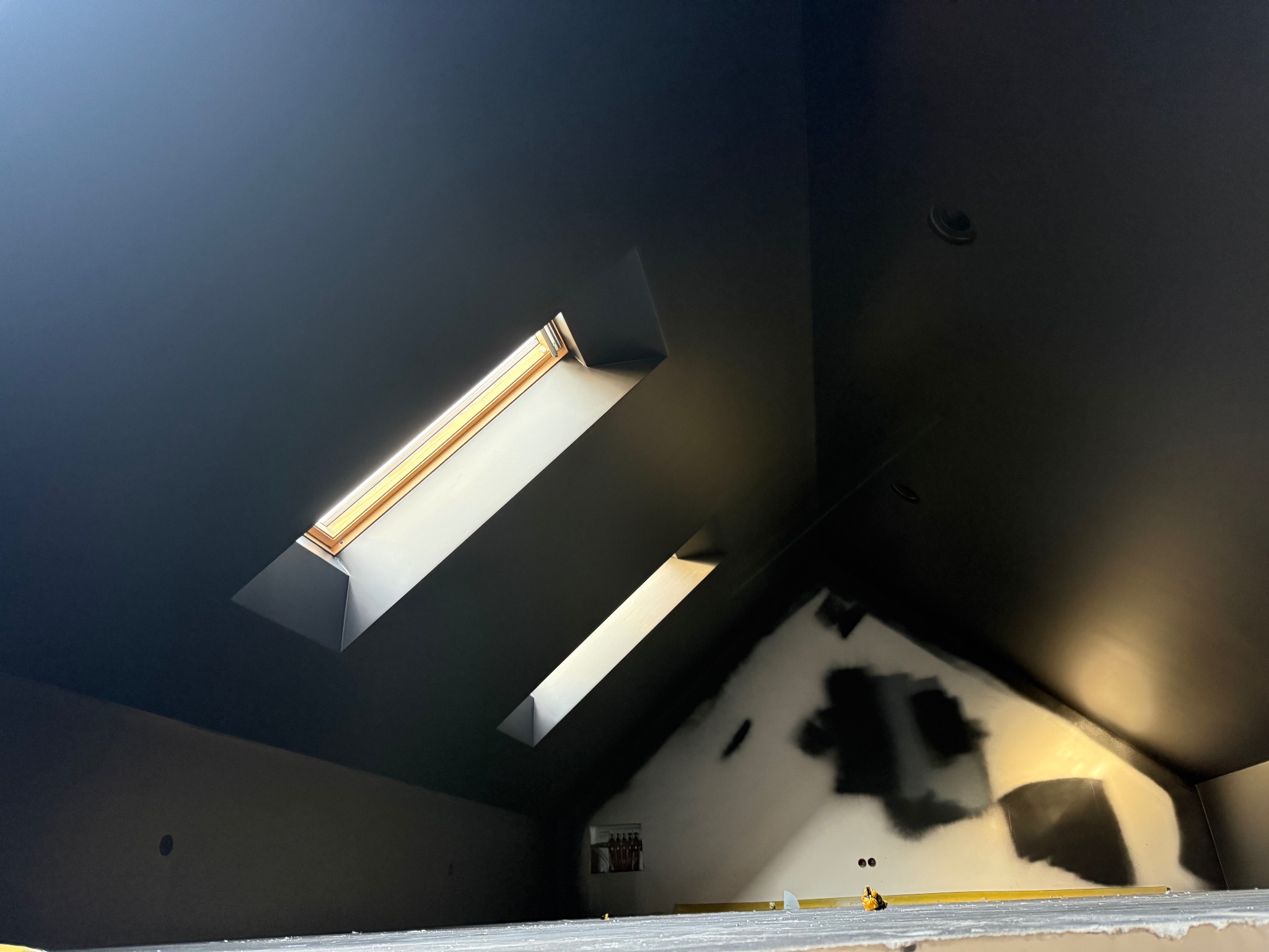 Poddasze po malowaniu na czarno, dwa okna dachowe wpuszczają światło. Fragment ściany z niedomalowanymi plamami. Widok z dołu, perspektywa.