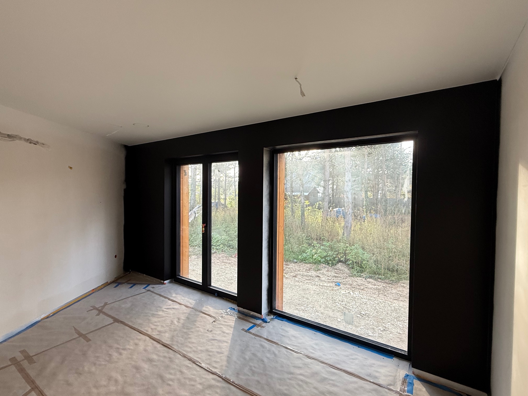 Pokój po malowaniu ścian na czarno, z zabezpieczoną folią podłogą i widokiem na zewnątrz przez duże okna. Ściany w trakcie prac wykończeniowych.