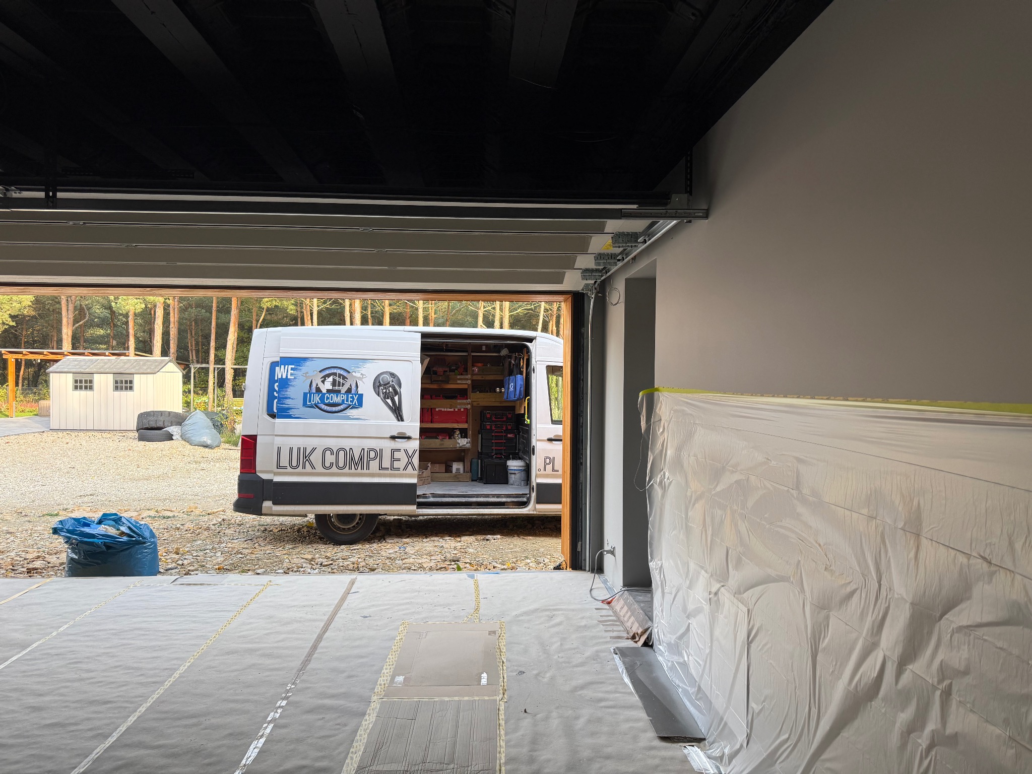 Wnętrze garażu w trakcie prac wykończeniowych: podłoga zabezpieczona folią, ściana osłonięta, na zewnątrz widoczny bus firmy LUK COMPLEX z otwartymi drzwiami.