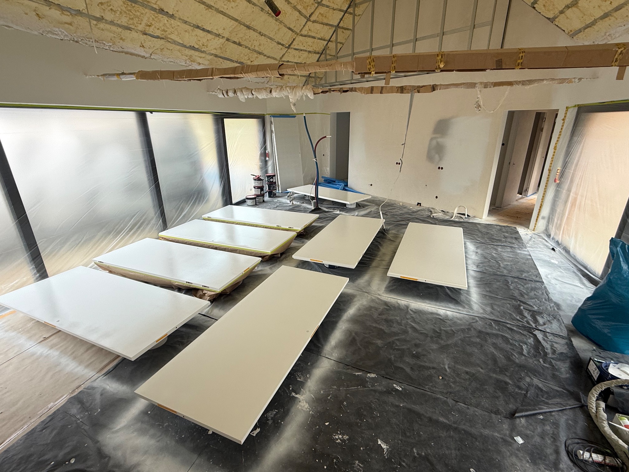 Białe, świeżo pomalowane drzwi ułożone na folii ochronnej w trakcie prac wykończeniowych wnętrza. Widoczne zabezpieczone okna i sufit z ociepleniem.