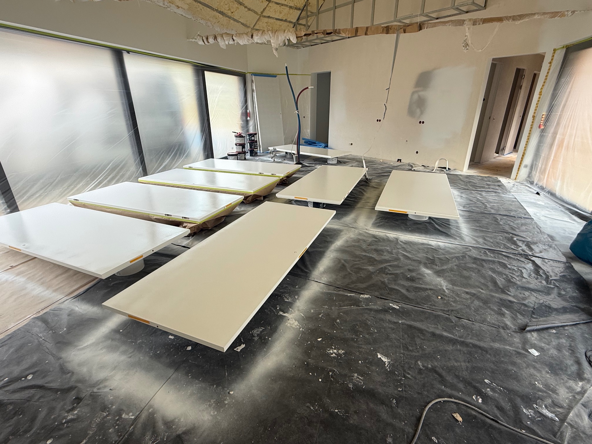 Malowanie drzwi w trakcie realizacji. Białe skrzydła drzwiowe leżą na folii ochronnej w pomieszczeniu w trakcie remontu. Widoczne puszki z farbą i narzędzia malarskie.