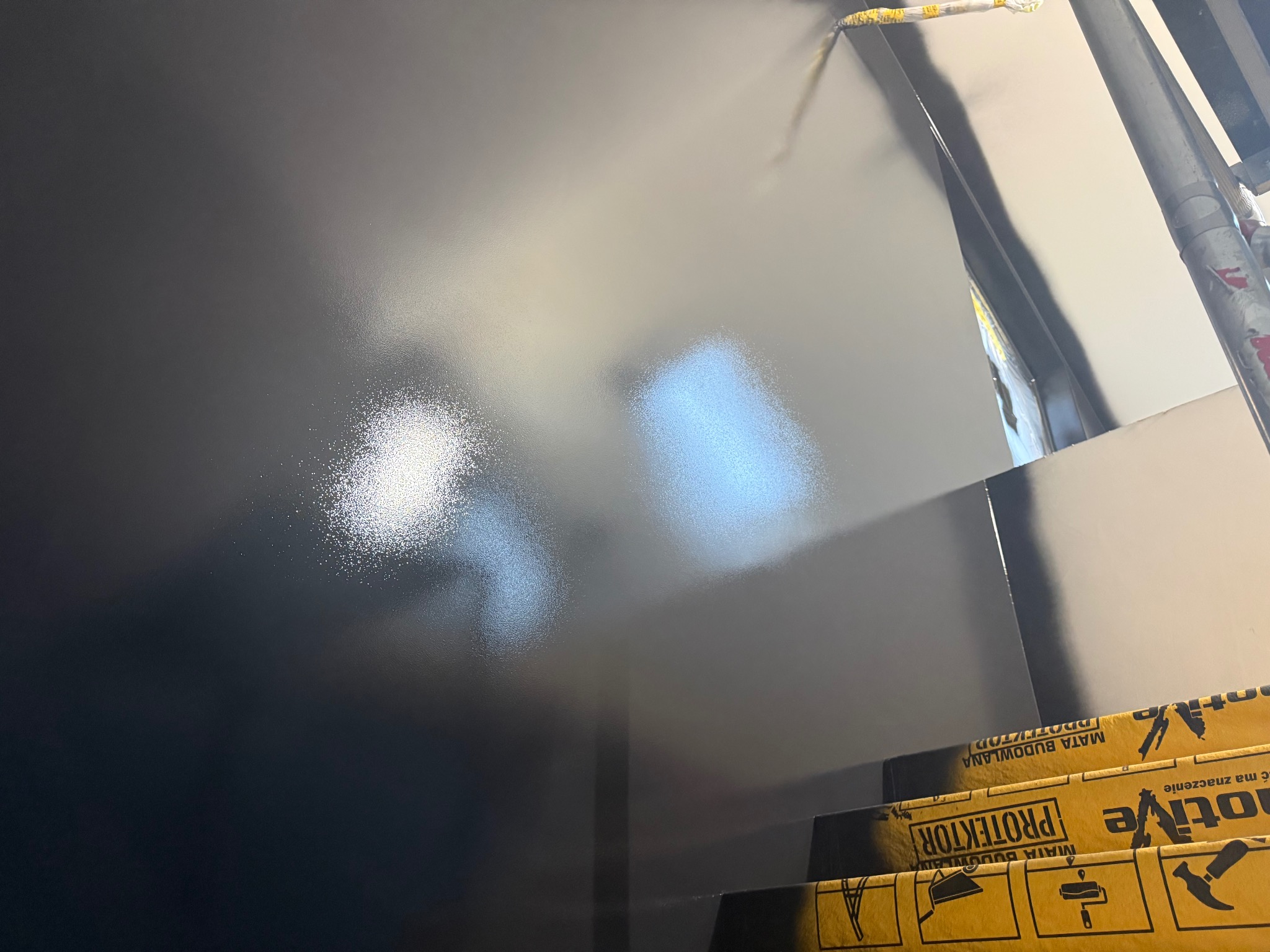 Świeżo pomalowana, gładka ściana w odcieniach szarości, z widocznym wałkiem malarskim u góry i zabezpieczeniem podłogi. Minimalistyczne ujęcie z bliska.