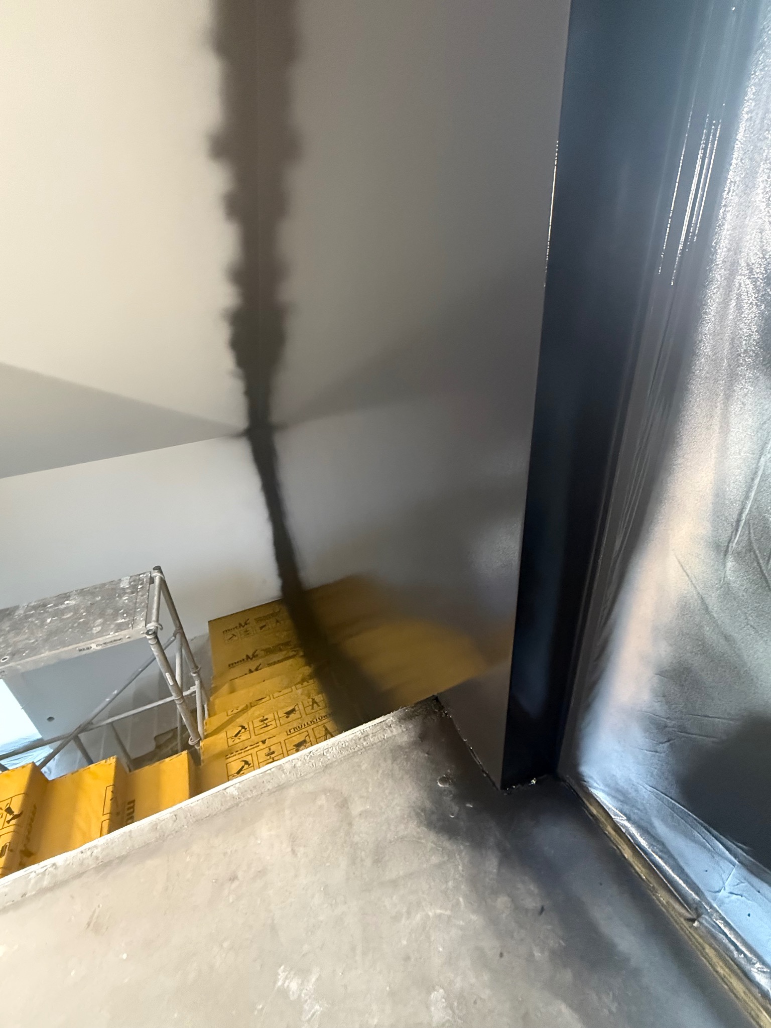 Przygotowanie do malowania: zabezpieczona folią framuga okienna, ściana z nałożoną warstwą farby, widoczne rusztowanie i kartony. Ujęcie z dołu.