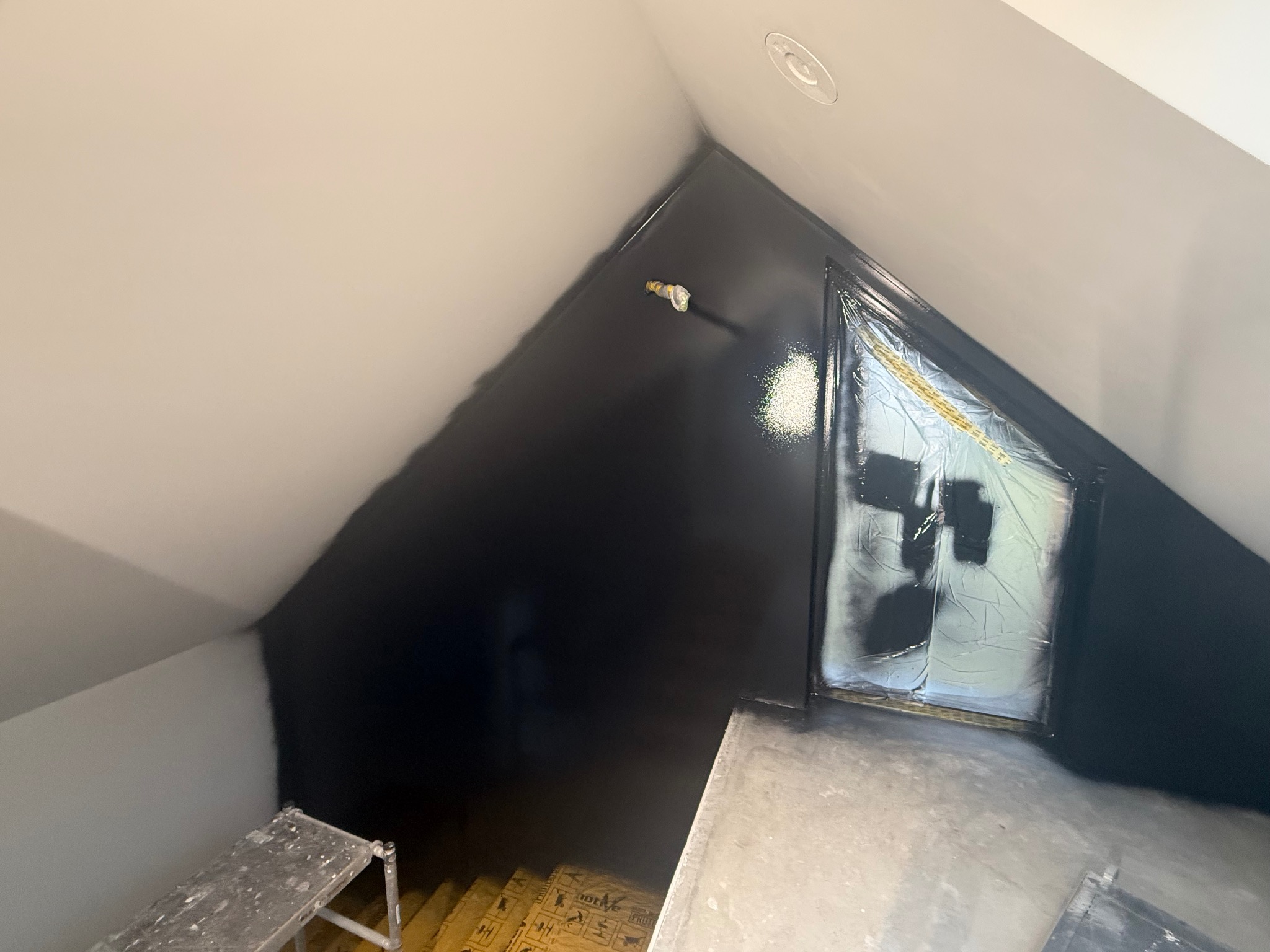 Ściana w skosie, częściowo pomalowana na czarno, zabezpieczone okno folią malarską. Widoczny fragment drabiny malarskiej i podłogi. Prace malarskie w trakcie realizacji.