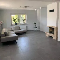 Jasnoszary salon po remoncie z narożną sofą, marmurowym kominkiem i roślinami doniczkowymi na parapecie.