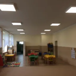 Sala przedszkolna w Czudcu z nowym oświetleniem: rzędy kwadratowych lamp LED w białych oprawach, równomiernie rozmieszczonych na suficie, oświetlających kolorowe stoliki i krzesła dla dzieci.