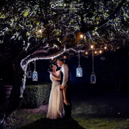 Para młoda w objęciach pod rozświetlonym drzewem z lampionami, ona w białej sukni z koronkowymi rękawami, on w ciemnym garniturze, nocna sceneria.