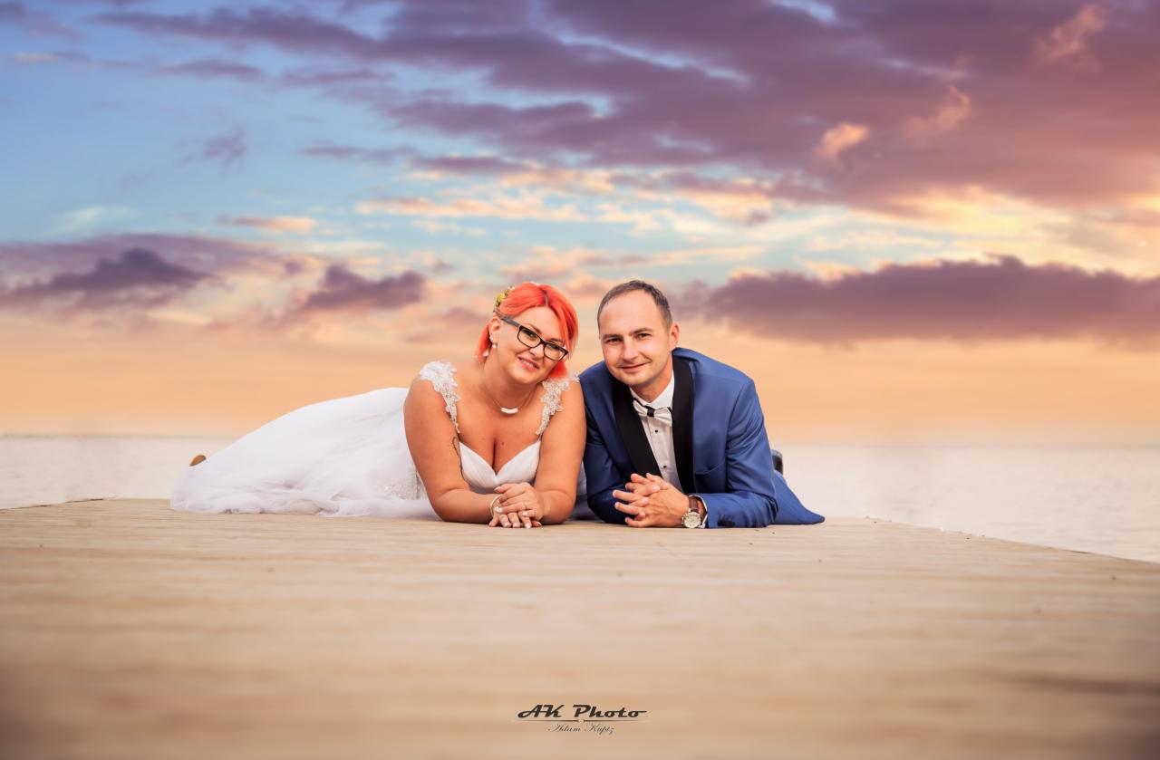 Para młoda leżąca na drewnianym pomoście z widokiem na zachodzące słońce i morze; panna młoda z rudymi włosami i okularami, pan młody w niebieskim smokingu.