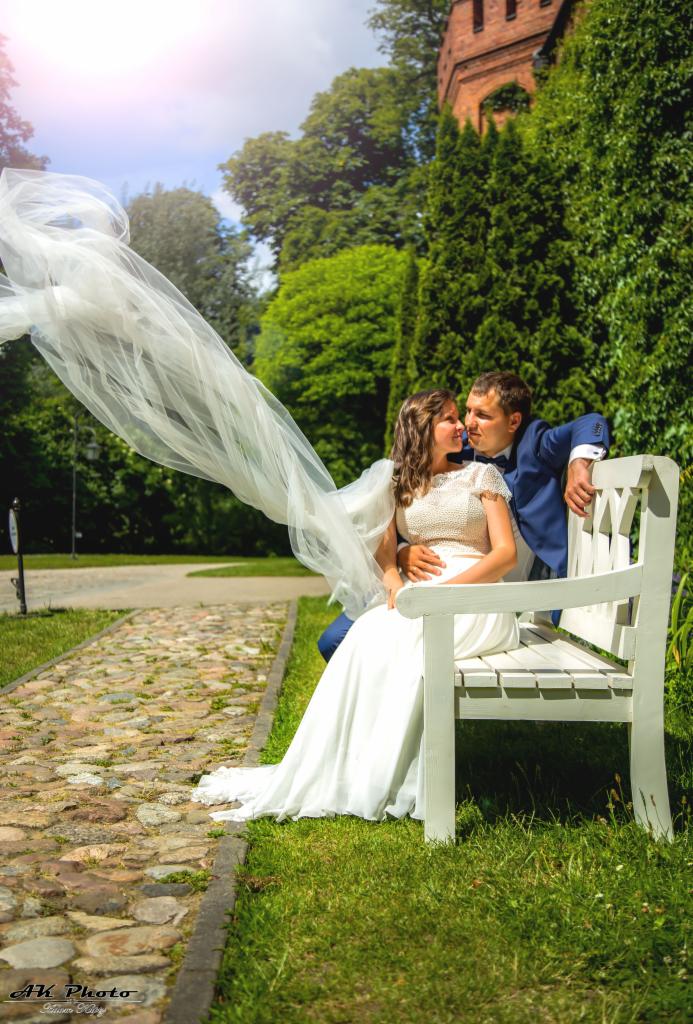 Para młoda siedzi na białej ławce w parku, welon powiewa na wietrze, w tle ceglana wieża.