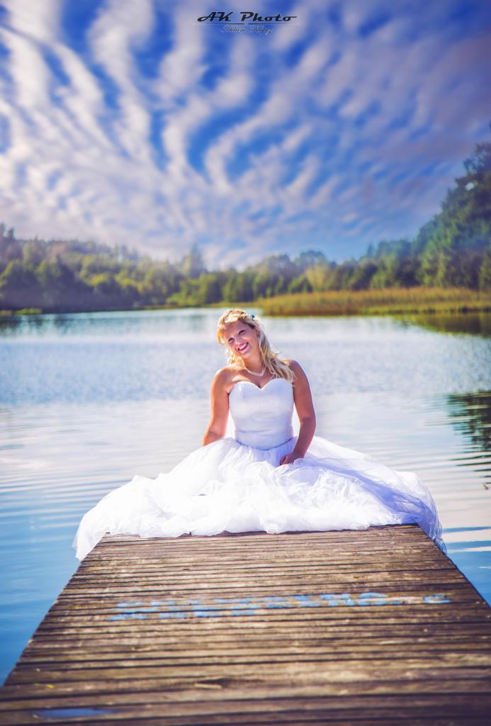 Uśmiechnięta panna młoda w białej sukni siedzi na drewnianym pomoście nad jeziorem, w tle las i błękitne niebo z chmurami.