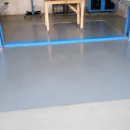 Dwukolumnowy podnośnik samochodowy z niebieskimi słupami na świeżo pomalowanej, jasnoszarej posadzce w warsztacie. W tle widoczne narzędzia i stół warsztatowy.