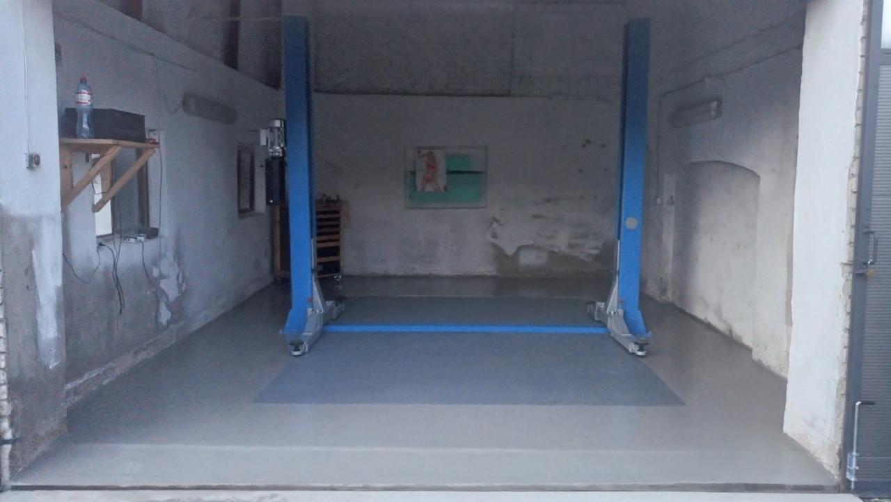 Pusty warsztat samochodowy z podnośnikiem dwukolumnowym w kolorze niebieskim, szarym betonowym podłożem i ścianami z widocznymi śladami napraw.