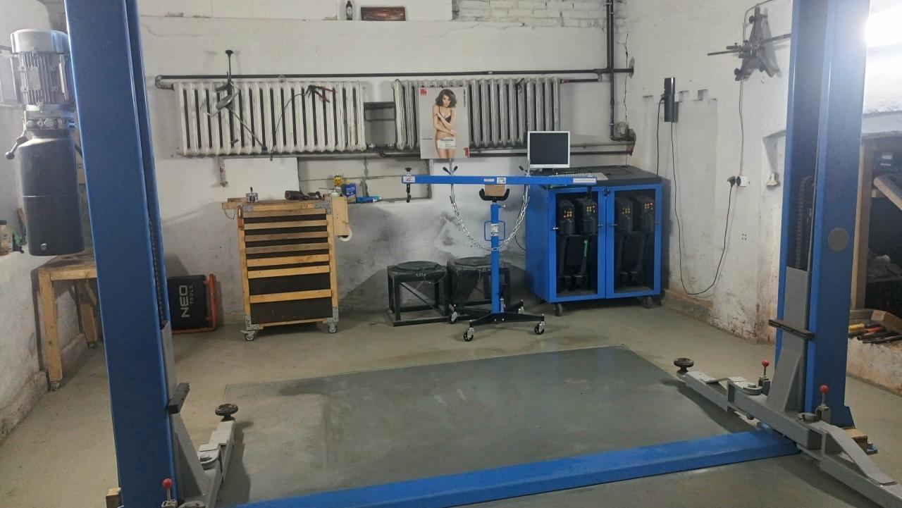 Dwukolumnowy podnośnik samochodowy w warsztacie z widocznym w tle wózkiem narzędziowym, podnośnikiem silnika i szafką z komputerem.