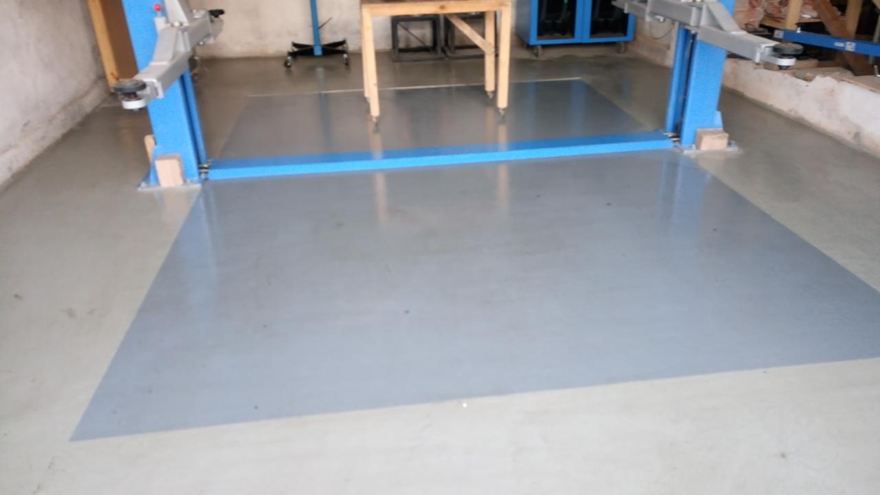 Dwukolumnowy podnośnik samochodowy z niebieskimi słupami na świeżo pomalowanej, jasnoszarej posadzce w warsztacie. W tle widoczne narzędzia i stół warsztatowy.