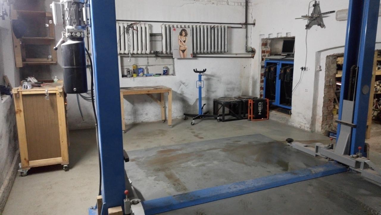 Wnętrze warsztatu samochodowego z podnośnikiem dwukolumnowym, widoczne narzędzia, stół roboczy i plakat z kobietą na ścianie.