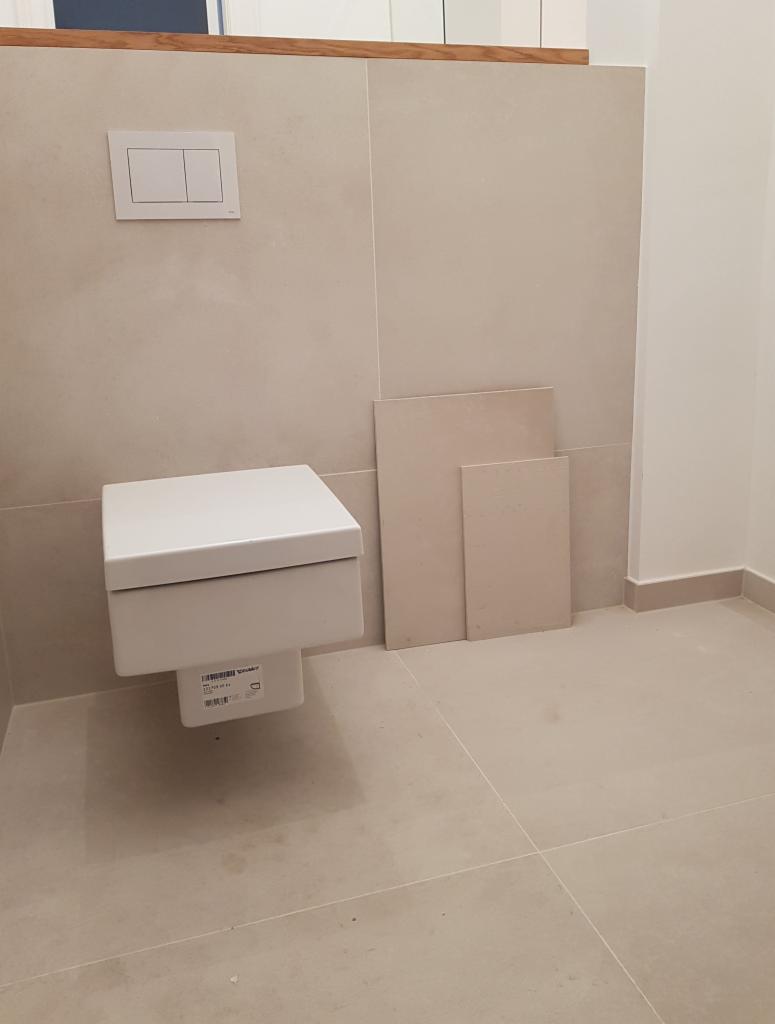 Wisząca toaleta z ukrytym zbiornikiem, zamontowana na ścianie wyłożonej beżowymi płytkami, obok leżą dwie dodatkowe płytki. Widoczny minimalistyczny przycisk spłuczki.