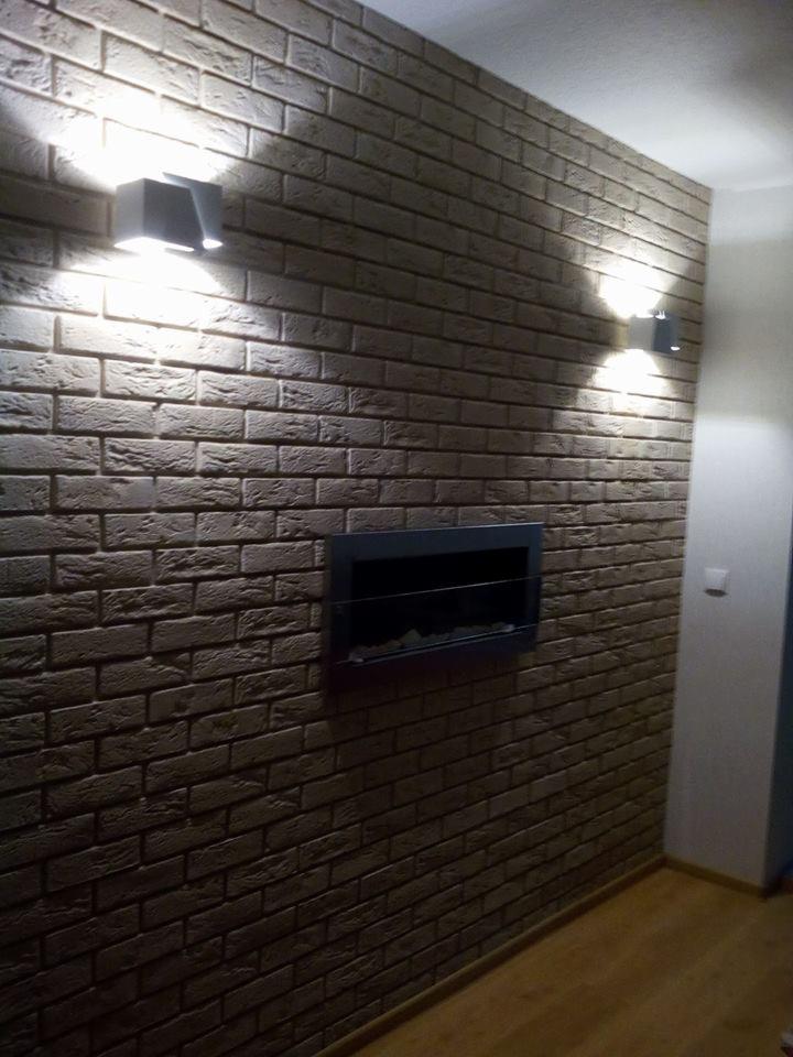 Ściana wykończona imitacją cegły w odcieniach szarości i beżu, oświetlona dwoma nowoczesnymi kinkietami, z wbudowanym biokominkiem.