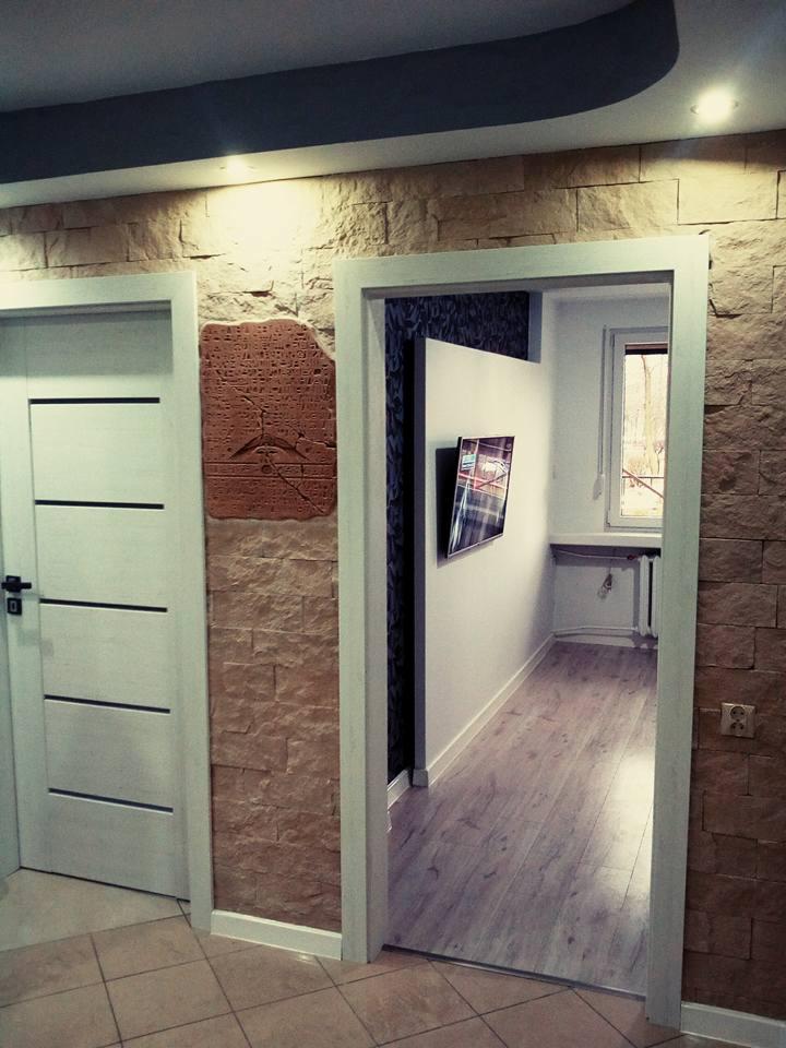 Wnętrze z dekoracyjną ścianą z kamienia, białe drzwi z poziomymi frezowaniami i widok na pokój z panelami podłogowymi i telewizorem na ścianie.