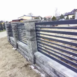 Fragment nowo postawionego ogrodzenia z szarego kamienia i metalowych, poziomych przęseł, widoczny na tle placu budowy z domami w tle.