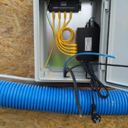 SNT - Otwarta skrzynka elektryczna z zamontowanym zabezpieczeniem AXON PoE Multi NetProtector HSK Data, widoczne żółte kable Ethernet, niebieska rura karbowana i przewody zasilające na tle płyty OSB.