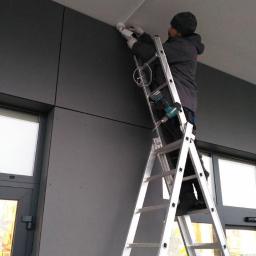 SNT - Mężczyzna w czapce i rękawicach montuje kamerę monitoringu na suficie, stojąc na aluminiowej drabinie, z wiertarką przymocowaną do drabiny.