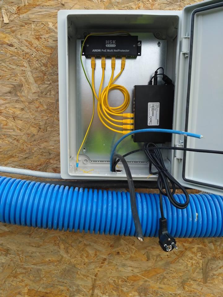 Otwarta skrzynka elektryczna z zamontowanym zabezpieczeniem AXON PoE Multi NetProtector HSK Data, widoczne żółte kable Ethernet, niebieska rura karbowana i przewody zasilające na tle płyty OSB.